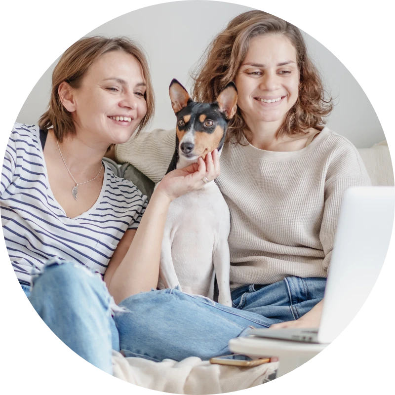 Two women sit on a couch and pet a dog.