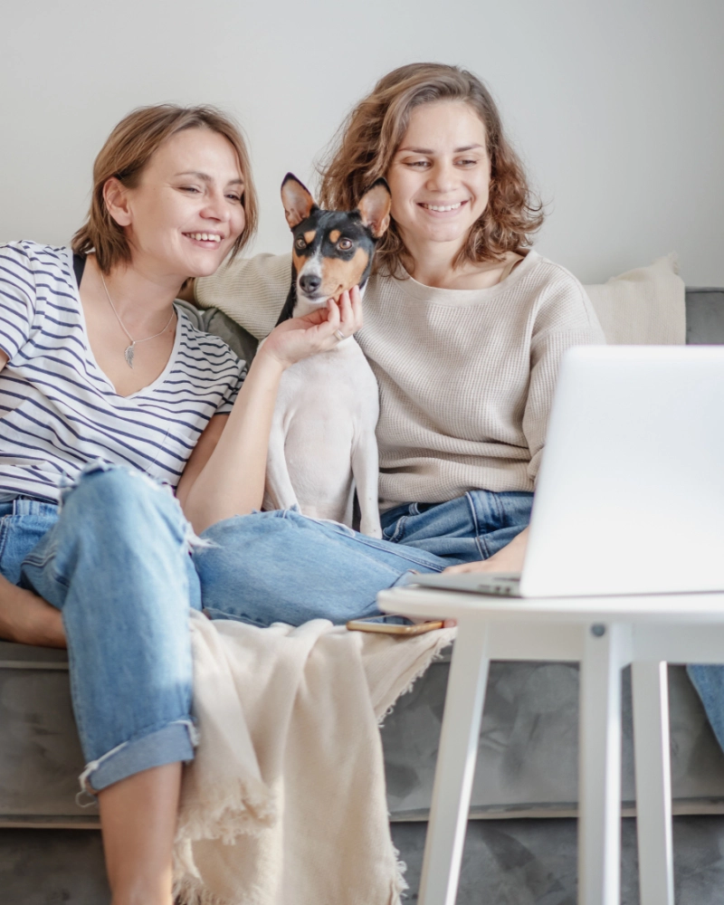 Two women sit on the couch with a dog, looking at a laptop.