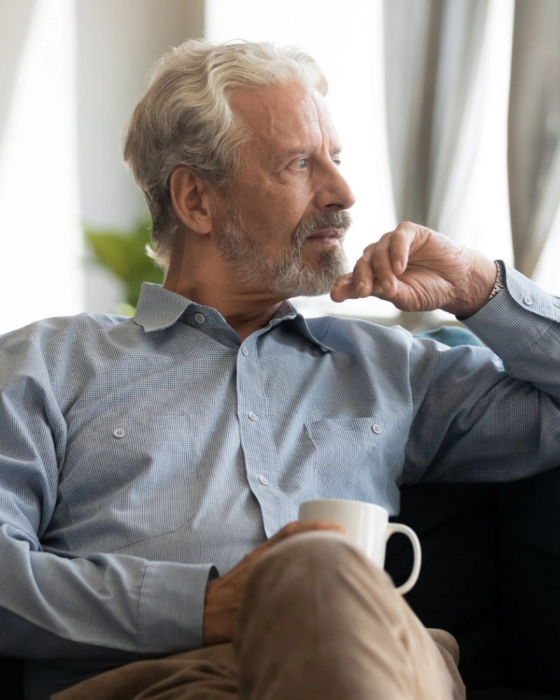 An older man sits on his couch and looks out the window.