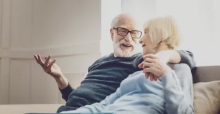 An older couple sitting on a couch, the man's arm around the woman.